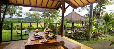 A wooden table with food and drinks in a covered outdoor area with a view of a garden and a thatched-roof structure.