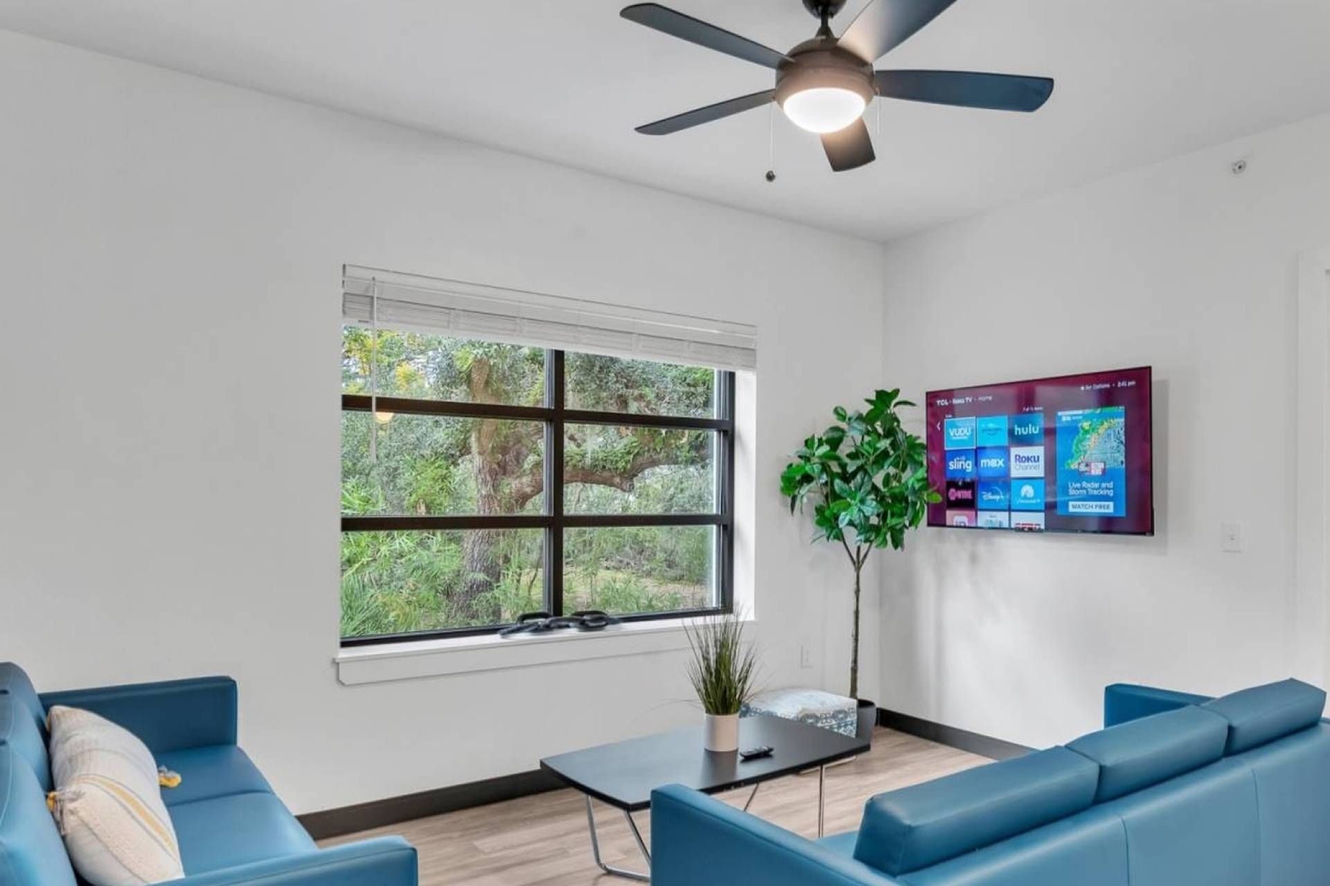 A modern living room with a large window, a ceiling fan, a flat-screen TV, and blue sofas.