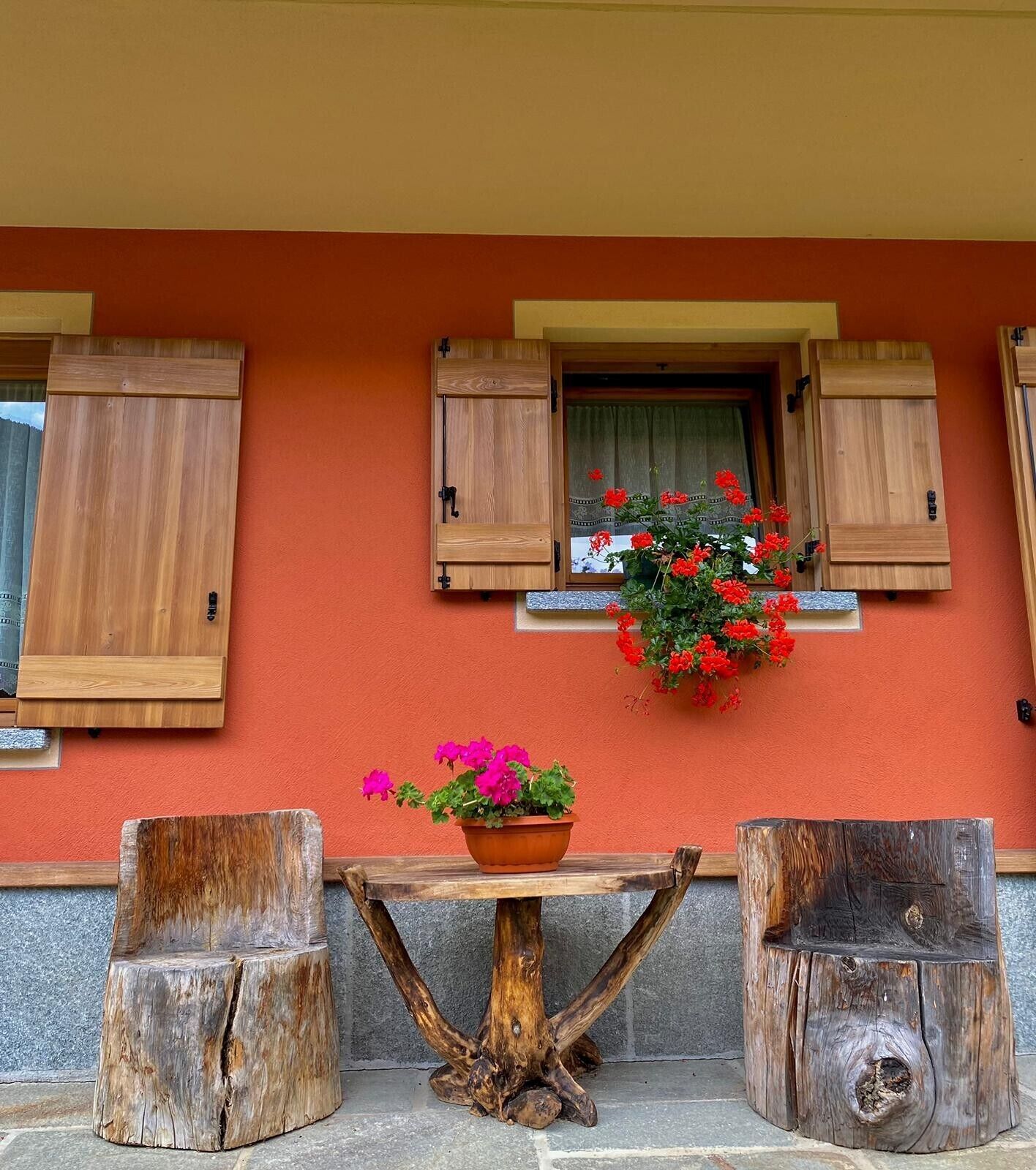 A wooden table with a potted plant and two wooden stools against a wall with a window and shutters.