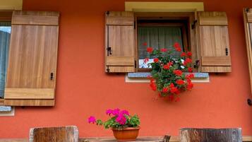 A wooden table with a potted plant and two wooden stools against a wall with a window and shutters.