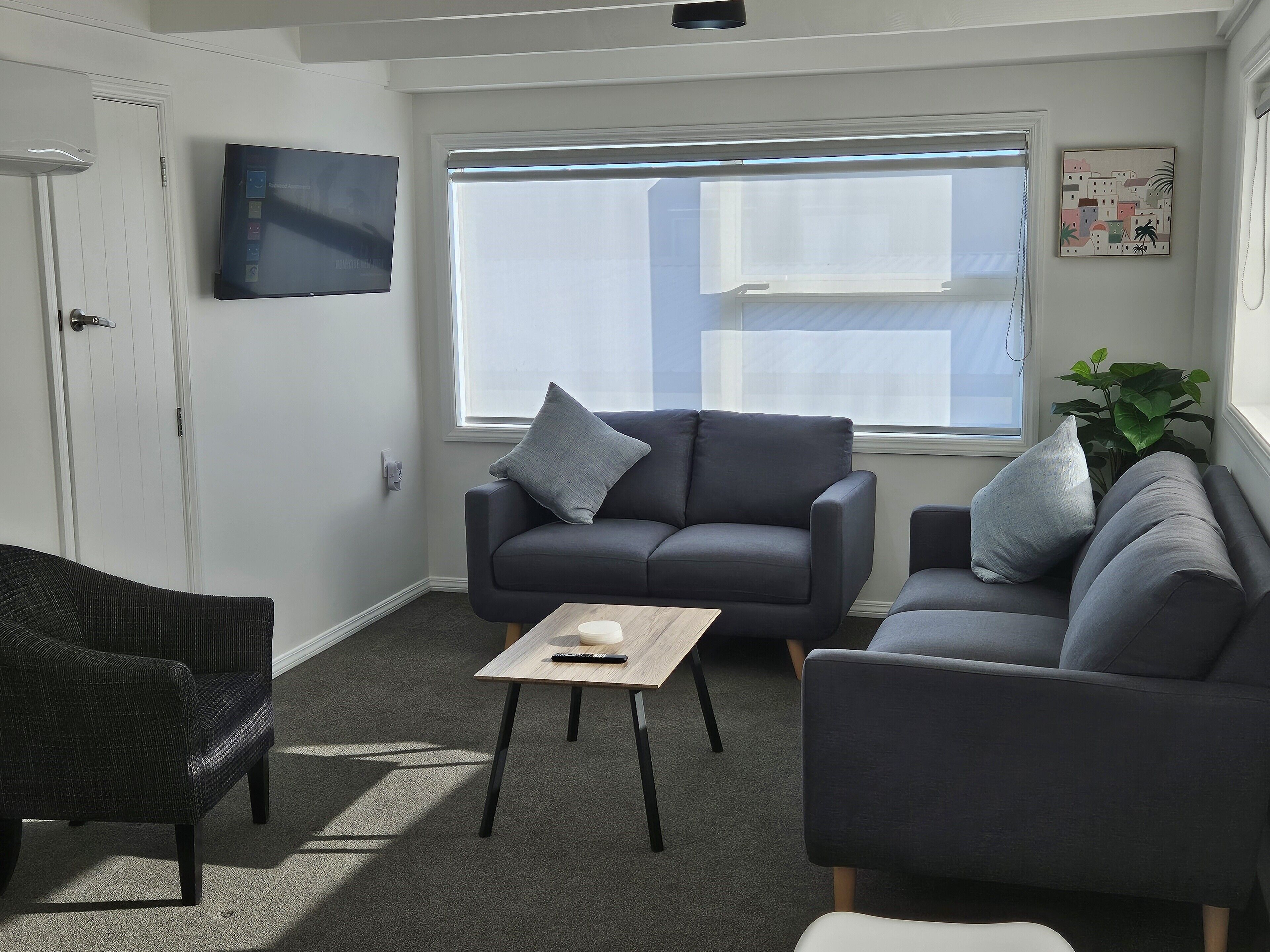 Una sala de estar con un sofá gris, una mesita, una silla de mimbre y un ventanal con persianas.
