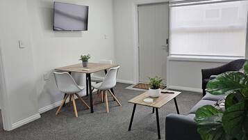A modern living room with a flat-screen TV mounted on the wall, a dining table with chairs, a coffee table, and a large potted plant.