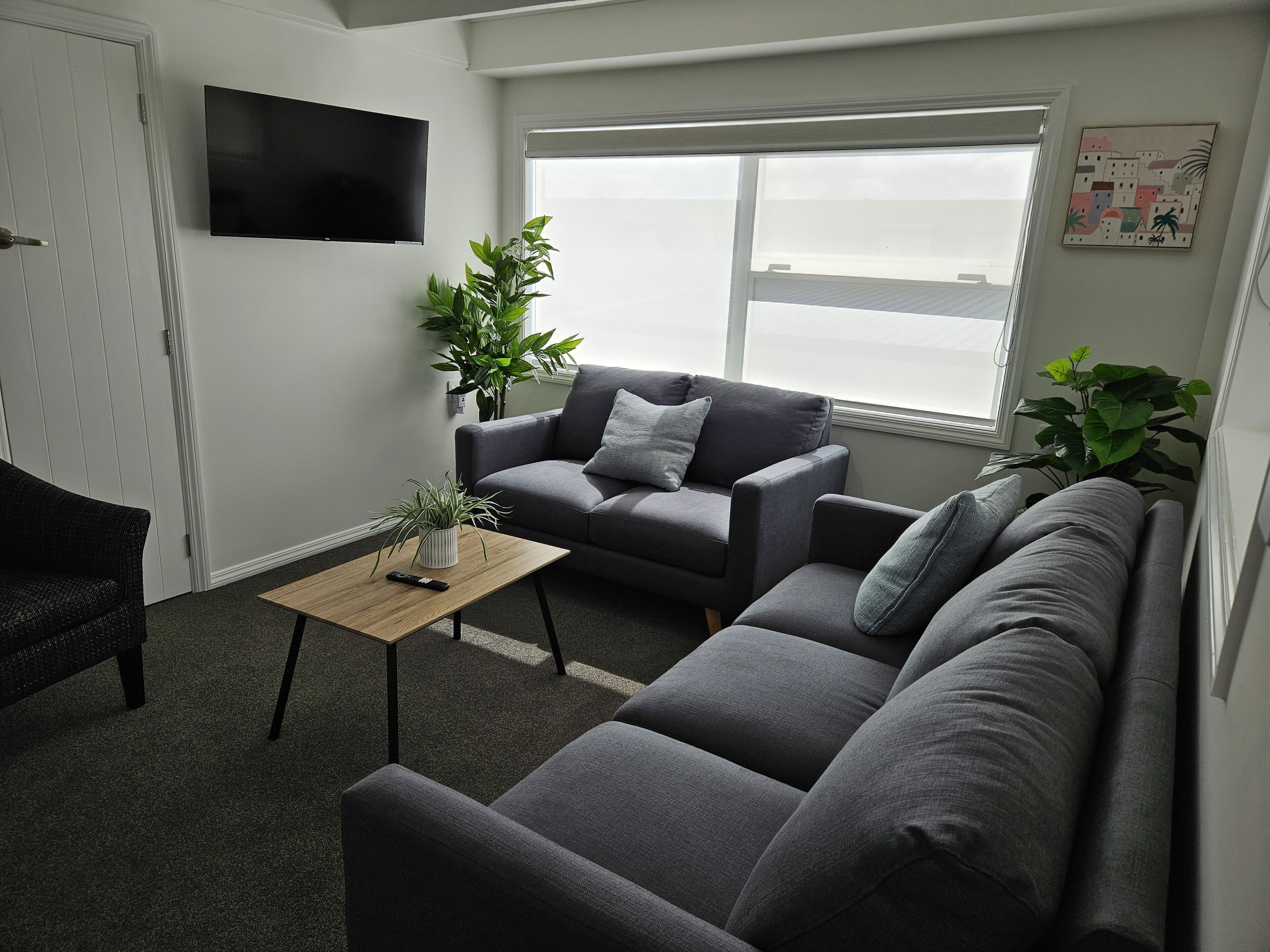 Een moderne woonkamer met een grijze zithoek, een houten salontafel, een aan de muur bevestigde televisie en een groot raam dat voor natuurlijk licht zorgt.