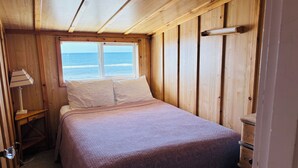 Un dormitorio de cabaña de madera con una cama, una mesita de noche, una lámpara y una ventana con vista al océano.