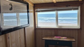 Una habitación con paredes de madera, una partitura enmarcada, una ventana con vistas a la playa y una pequeña mesa con un libro.
