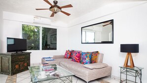 A living room with a sofa, colorful cushions, a glass coffee table, a television, a ceiling fan, a large mirror, and a small table with a lamp. - Amazing ocean view from Estrellita de Mar 303 2BD Condo for rent in Amapas, Puer (Puerto Vallarta)