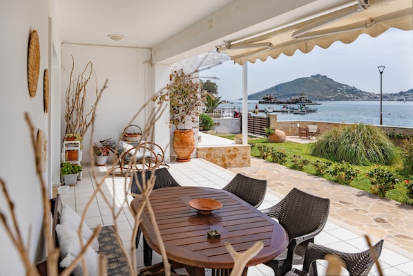 A covered patio with a table and chairs, overlooking a body of water with boats and a hill in the distance.