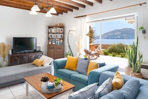 A living room with a sofa, coffee table, bookshelf, and a TV. There's a view of the sea and a balcony with plants.