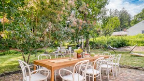 Un patio al aire libre con una mesa de madera y sillas blancas, rodeado de follaje y una hamaca.