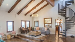 A spacious living room with a high ceiling, wooden beams, a staircase, and a view of the outdoors. - 20-Acre Retreat • Hot Tub + Panoramic Views + Pickleball Court | Slice of Heaven (Temecula)