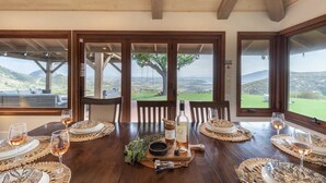 A dining table set for six with wine glasses, plates, and a bottle of wine. Large windows offer a view of a green landscape. - 20-Acre Retreat • Hot Tub + Panoramic Views + Pickleball Court | Slice of Heaven (Temecula)