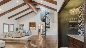 A spacious living area with a high ceiling and exposed beams, a modern staircase, and a kitchen with a herringbone tile backsplash. - 20-Acre Retreat • Hot Tub + Panoramic Views + Pickleball Court | Slice of Heaven (Temecula)