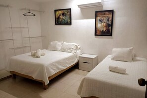 A bedroom with two single beds, white bedding, a small bedside table with a drawer, and a wall-mounted shelf.