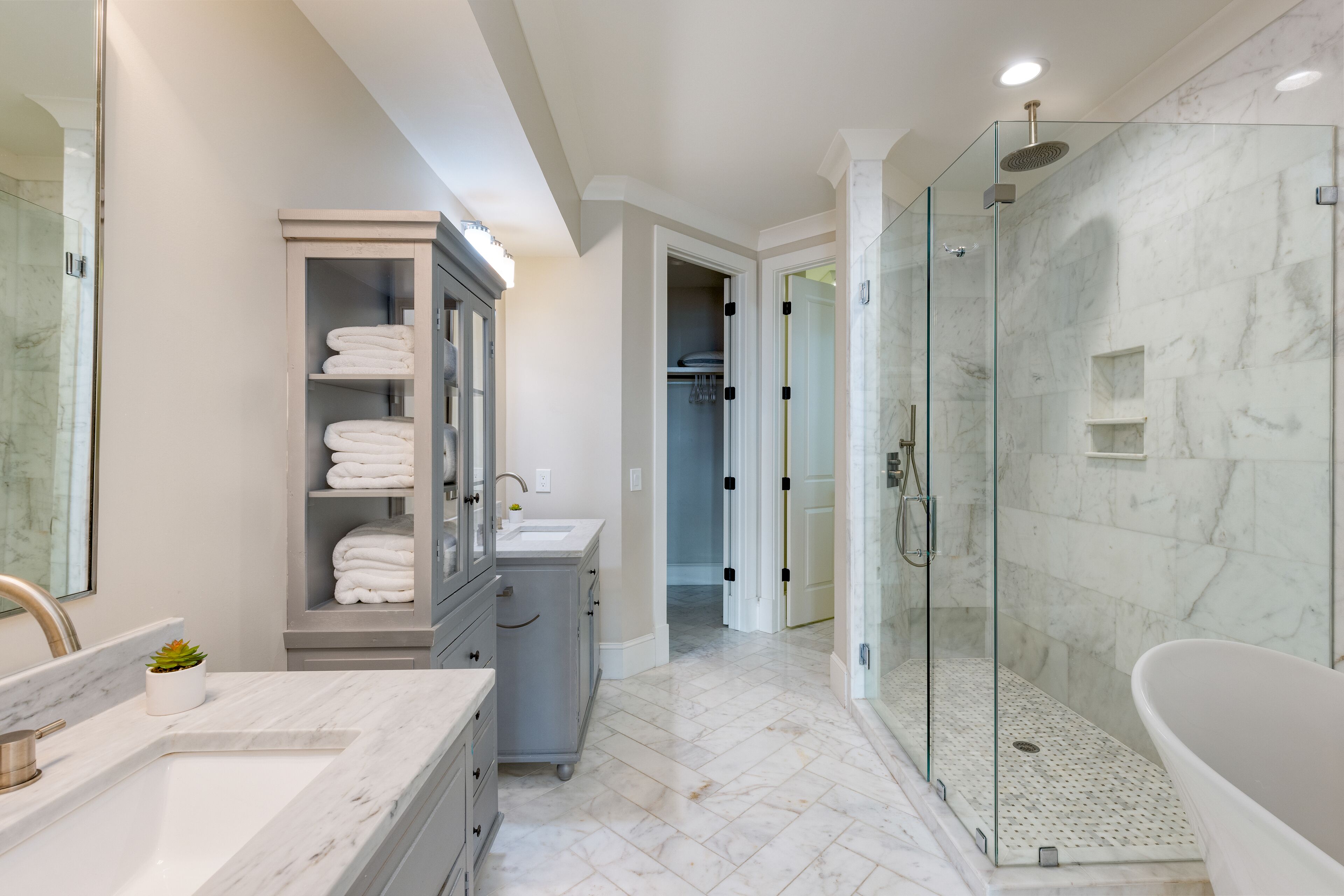 A modern bathroom with a glass-enclosed shower, a freestanding bathtub, a vanity with a sink, and a cabinet with folded towels.