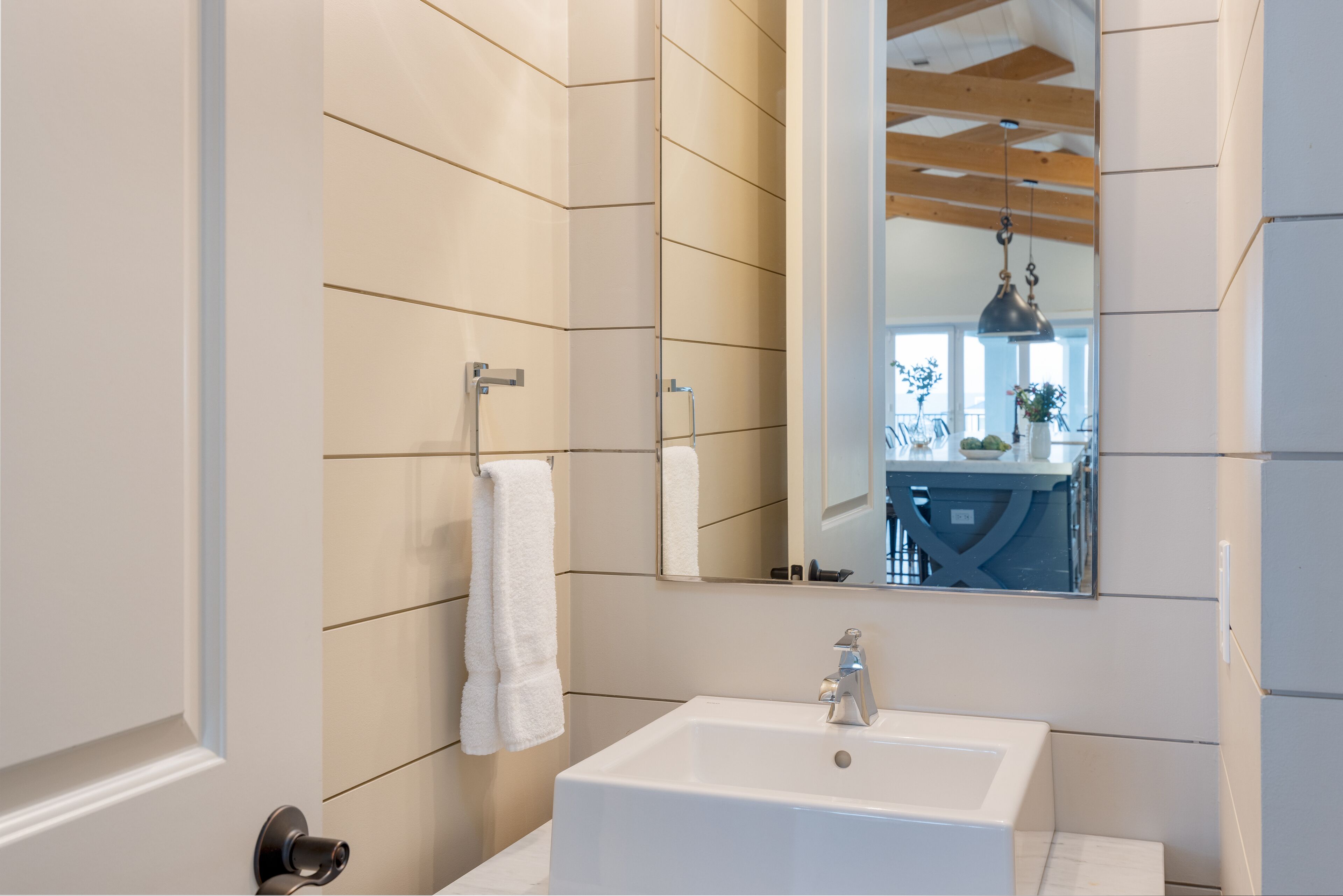 A bathroom with a white sink, a towel hanging on a rack, and a mirror reflecting a kitchen area.