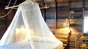 Ein Zimmer mit Holzboden, einem Bett unter einem Moskitonetz, einem Stuhl und einer Wandleuchte.