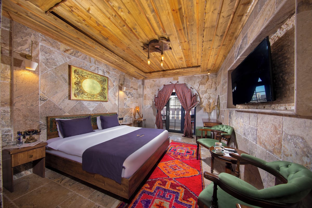 Quarto com parede de pedra, teto de madeira, cama com colcha roxa, poltrona verde e televisão de tela plana fixada na parede.
