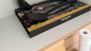 Ein Set Küchenutensilien auf einem Regal, darunter eine Bratpfanne mit Deckel und ein Pfannenwender-Set in einer Schachtel mit der Aufschrift "MICHELINO".