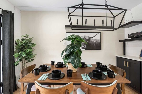 A dining area with a wooden table set for four, surrounded by chairs, a large potted plant, and a black pendant light.
