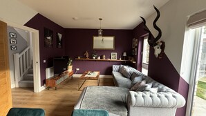 Una sala de estar con un sofá gris, sillones turquesa, una mesa de centro de madera y un televisor. Se ve una escalera a la izquierda y una ventana a la derecha.