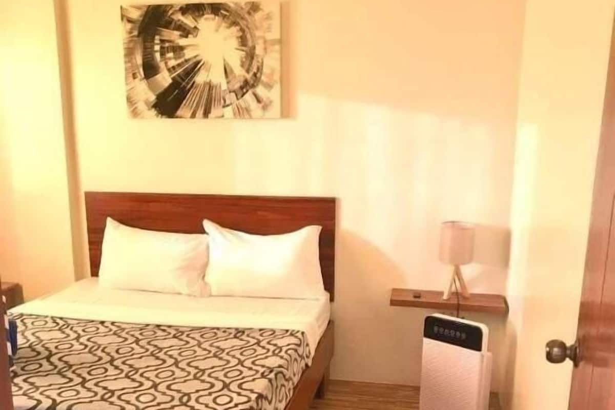 A hotel room with a wooden bed, patterned bedspread, a small bedside table with a lamp, and a wall-mounted artwork.