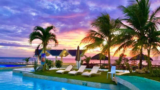 A poolside area with lounge chairs and palm trees, overlooking a beach at sunset. - Acapulco Resort & Suites (Toledo City)