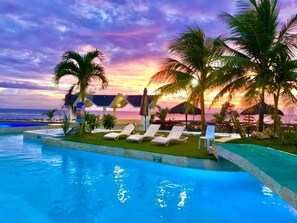 A poolside area with lounge chairs and palm trees, overlooking a beach at sunset. - Acapulco Resort & Suites (Toledo City)