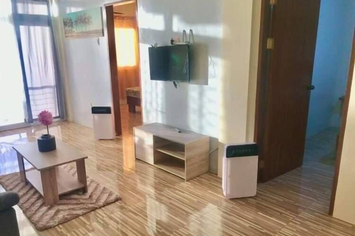A room with a wooden floor, a small table with a vase, a TV mounted on the wall, and a door leading to another room.