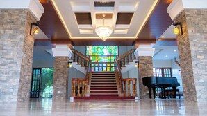 A grand staircase with a chandelier, leading to a piano, surrounded by stone pillars and a stained glass window. - Kasa Cailloux (Pasuquin)
