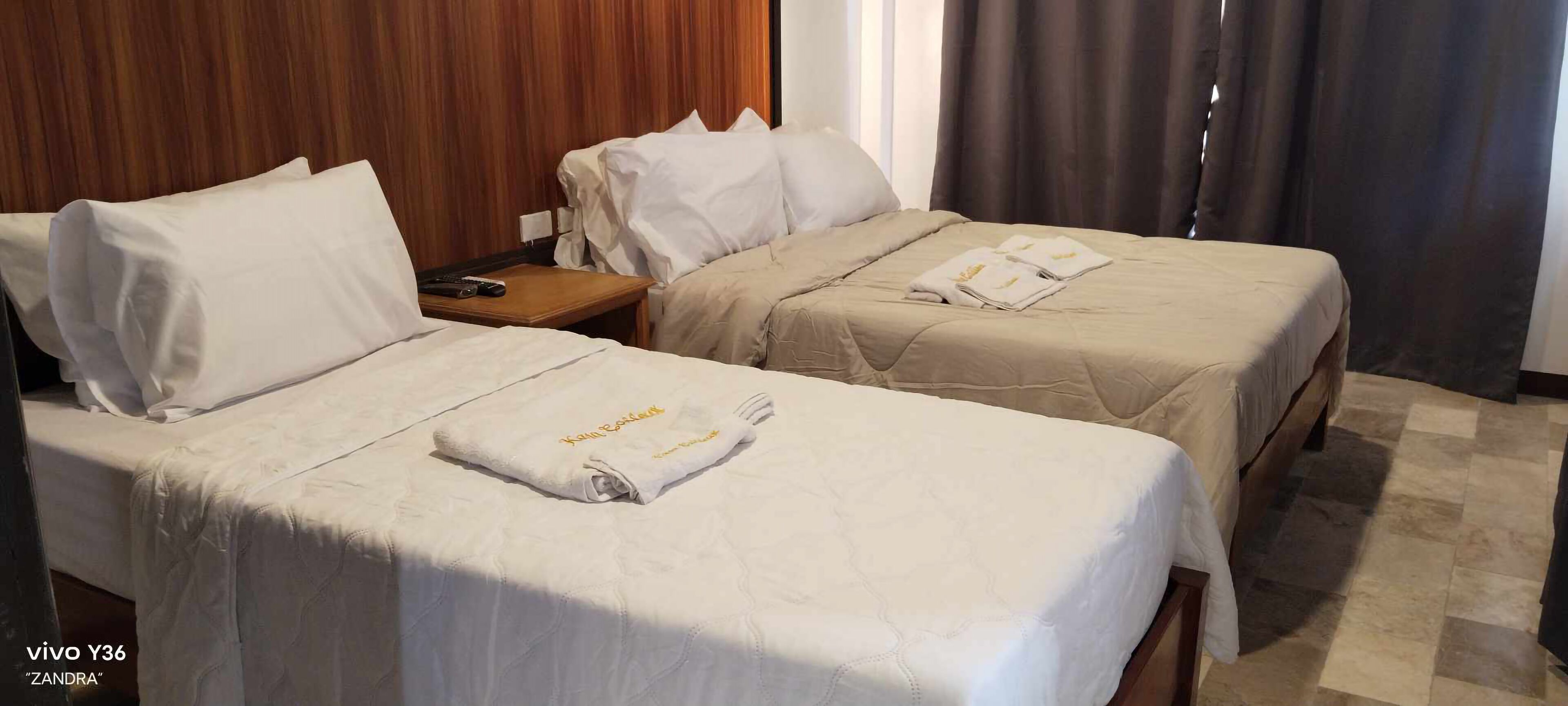 A hotel room with two beds, each with white linens and a beige blanket. There is a wooden headboard and a nightstand with a remote control.