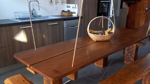 Uma mesa de jantar de madeira com bancos, suspensa por cordas, em uma cozinha com armários, uma máquina de lavar louça e um relógio na parede.