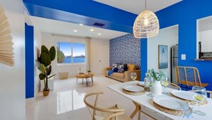 A modern living room with a blue accent wall, a dining area with a white table set for four, and a large window offering a view of the sea. - Modern Beachside: Restaurants, Pool (Hollywood)