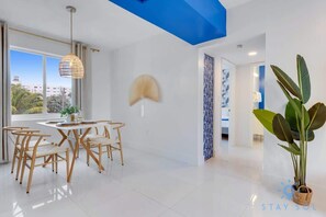 A modern dining area with a white table and chairs, a hanging light fixture, and a view of the cityscape through large windows. - Modern Beachside: Restaurants, Pool (Hollywood)