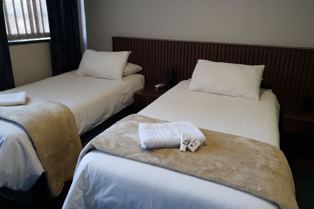 Une chambre d’hôtel avec deux lits simples, chacun recouvert d’une courtepointe blanche et d’un plaid beige. Un des lits est doté d’une tête de lit en bois, et l’autre est muni d’une table de chevet avec une lampe et un téléphone.