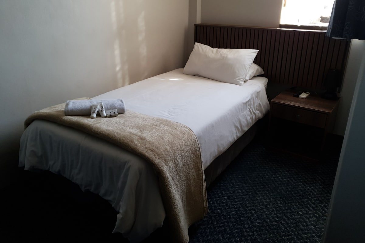 Une chambre d’hôtel avec un lit simple, une table de chevet avec une lampe et une fenêtre avec des rideaux.