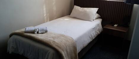 Ein Hotelzimmer mit einem Einzelbett, einem Nachttisch mit Lampe und einem Fenster mit Vorhängen.