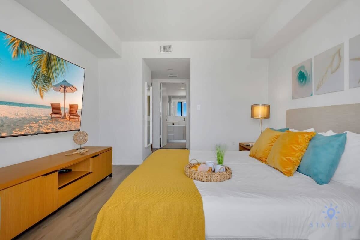 A hotel room with a bed, a TV on the wall, a wooden side table, a yellow throw blanket, and a basket with items on the bed.