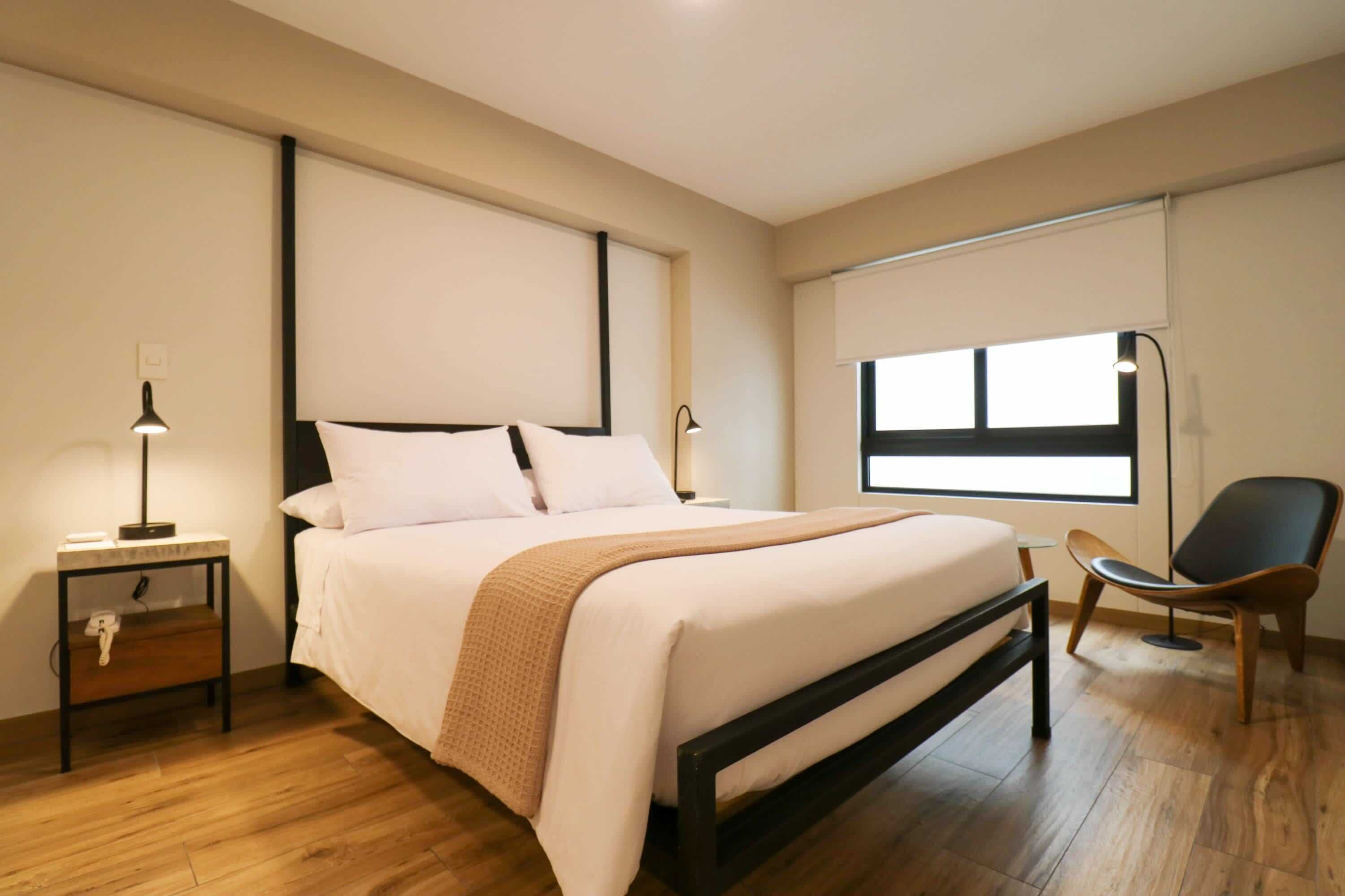 Une chambre d’hôtel moderne avec un grand lit, des tables de chevet avec des lampes, une chaise et une fenêtre avec des stores.
