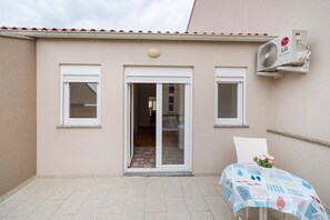 Un edificio con fachada color beige, un patio con baldosas, una puerta corrediza de vidrio y una unidad de aire acondicionado.