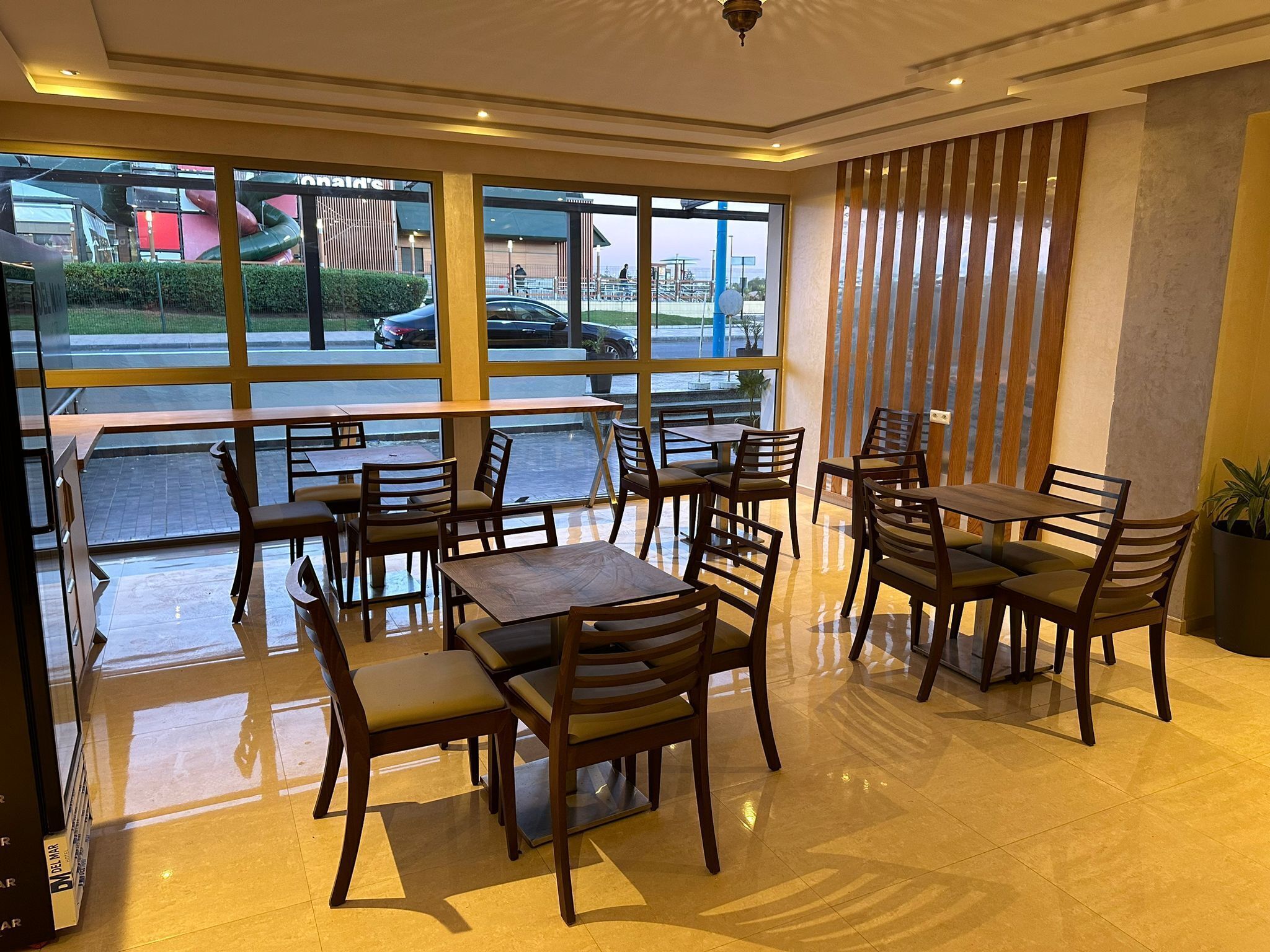A restaurant with wooden tables and chairs, large windows offering a view of the street and parked cars.