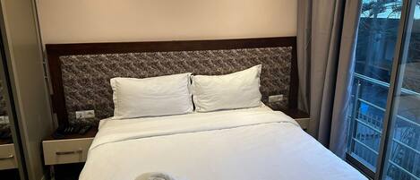 Une chambre d’hôtel avec un lit bien fait, une tête de lit ornée d’un motif et une fenêtre donnant sur des arbres.