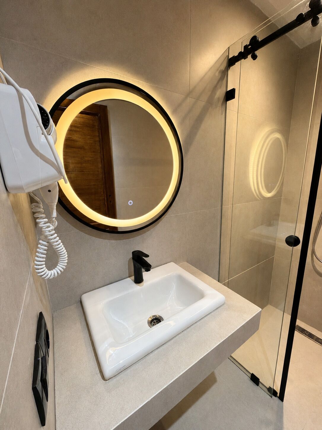 Ein modernes Badezimmer mit einem weißen Waschbecken, einem runden Spiegel mit Beleuchtung und einem an der Wand montierten Föhn.
