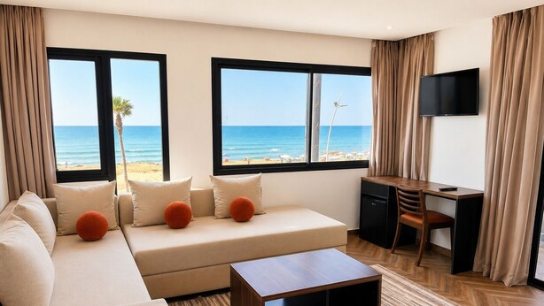 A modern hotel room with a sofa, a desk, a television, and large windows overlooking the beach. - Oceana Mazagan Suites Hotel  (El Jadida)