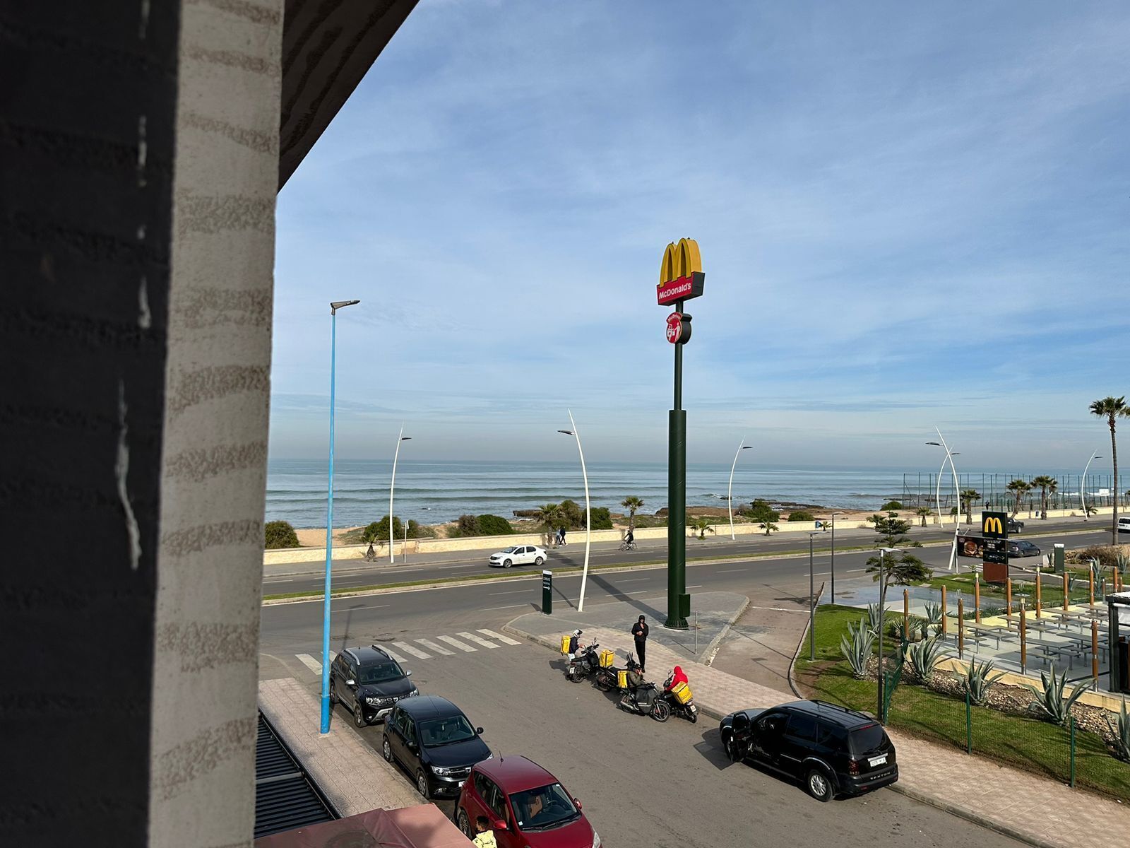A McDonald's sign with a view of the ocean and a parking lot with cars and motorcycles.