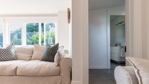 Ein Wohnzimmer mit einem beigen Sofa, gemusterten Kissen und Blick auf einen Garten durch große Fenster.