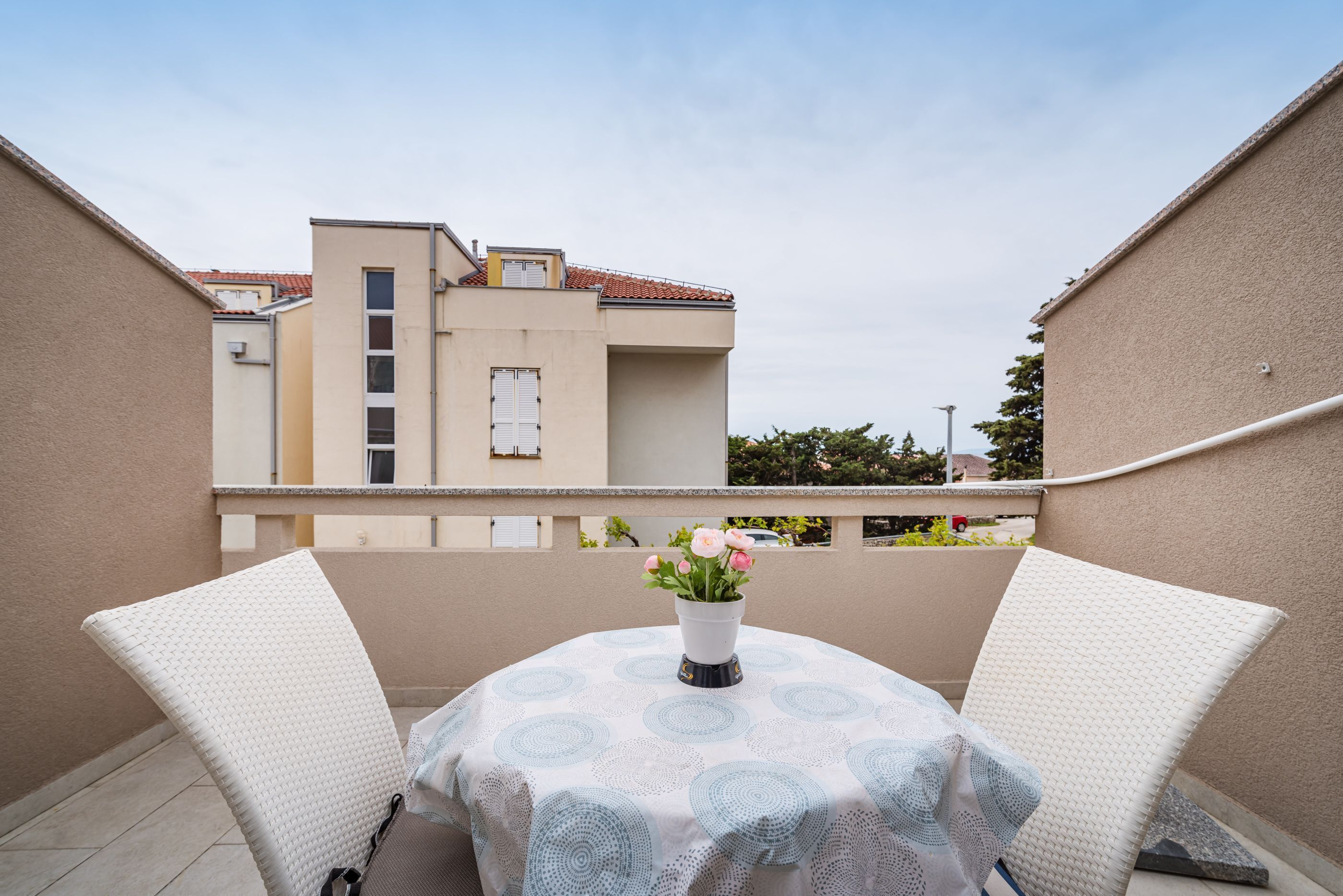 A balcony with two white wicker chairs, a table with a blue and white patterned tablecloth, and a vase with flowers.