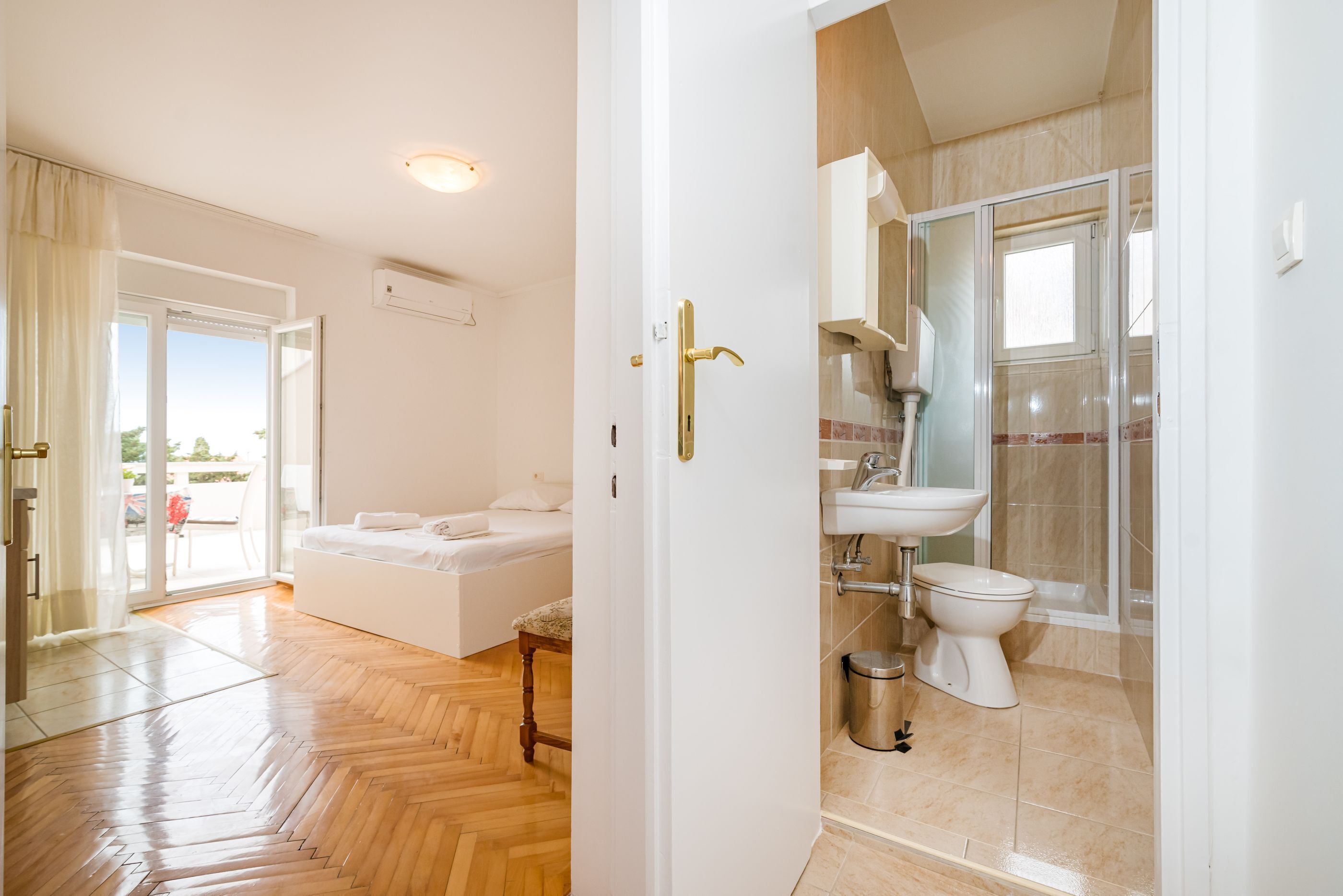 Une chambre d’hôtel avec un plancher en bois, un lit, une salle de bain avec un lavabo et des toilettes, et une porte-fenêtre coulissante donnant sur un balcon.