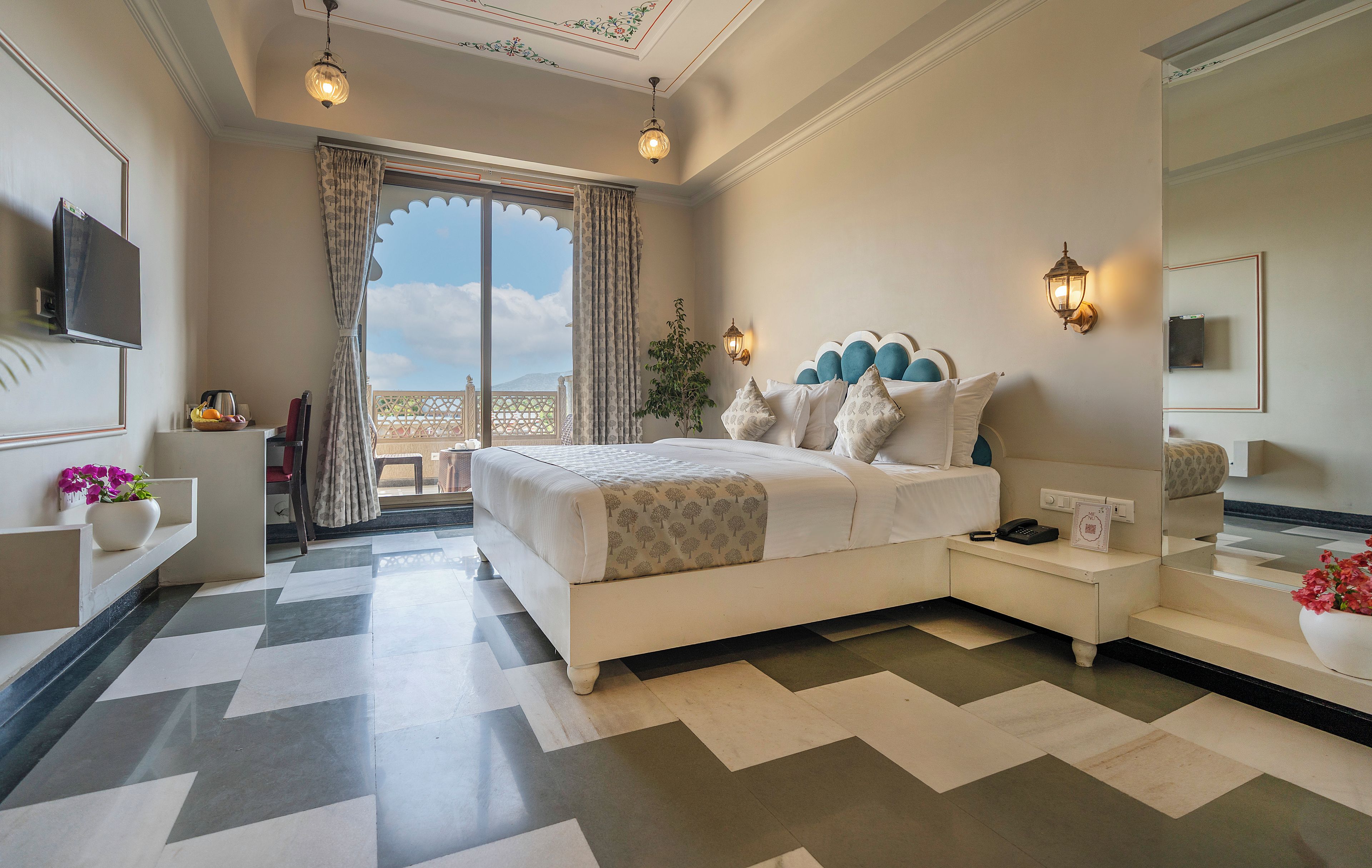 A hotel room with a bed, bedside table, lamp, and a view of the outdoors.