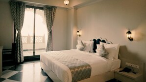 A hotel room with a bed, a desk, a chair, a nightstand, and a wall-mounted lamp. - The Biletha Bagh (Udaipur)