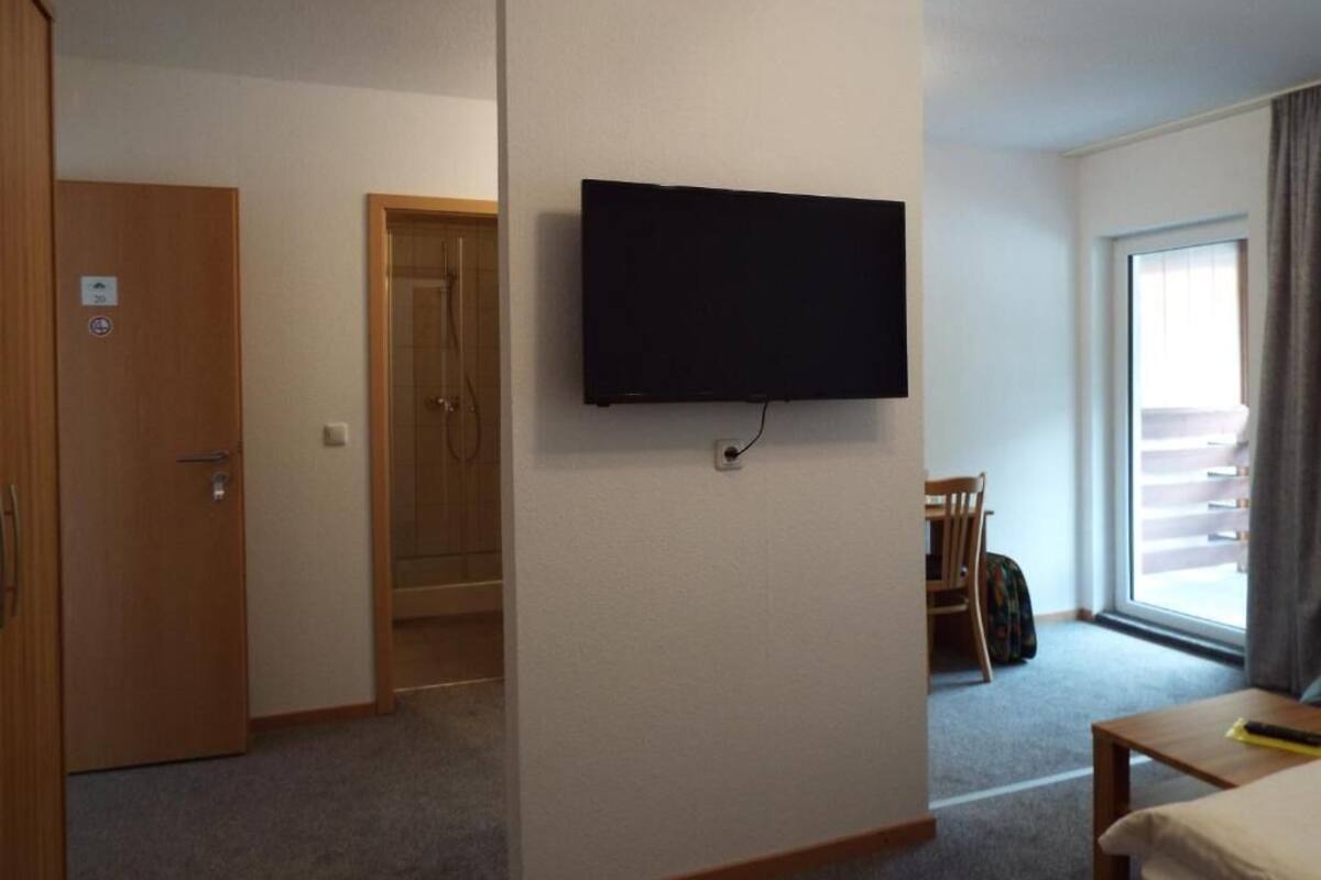 Habitación de hotel con televisor montado, puerta, zona de ducha y vistas al balcón exterior.